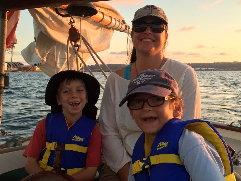 Family enjoying an intimate sailing experience on San Diego Bay aboard a historic Friendship sloop, with children wearing life jackets and smiling at sunset.