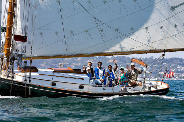 Group of six happy guests enjoying an intimate sailing experience aboard a historic Friendship sloop on San Diego Bay, with sails raised and scenic coastline in the background.